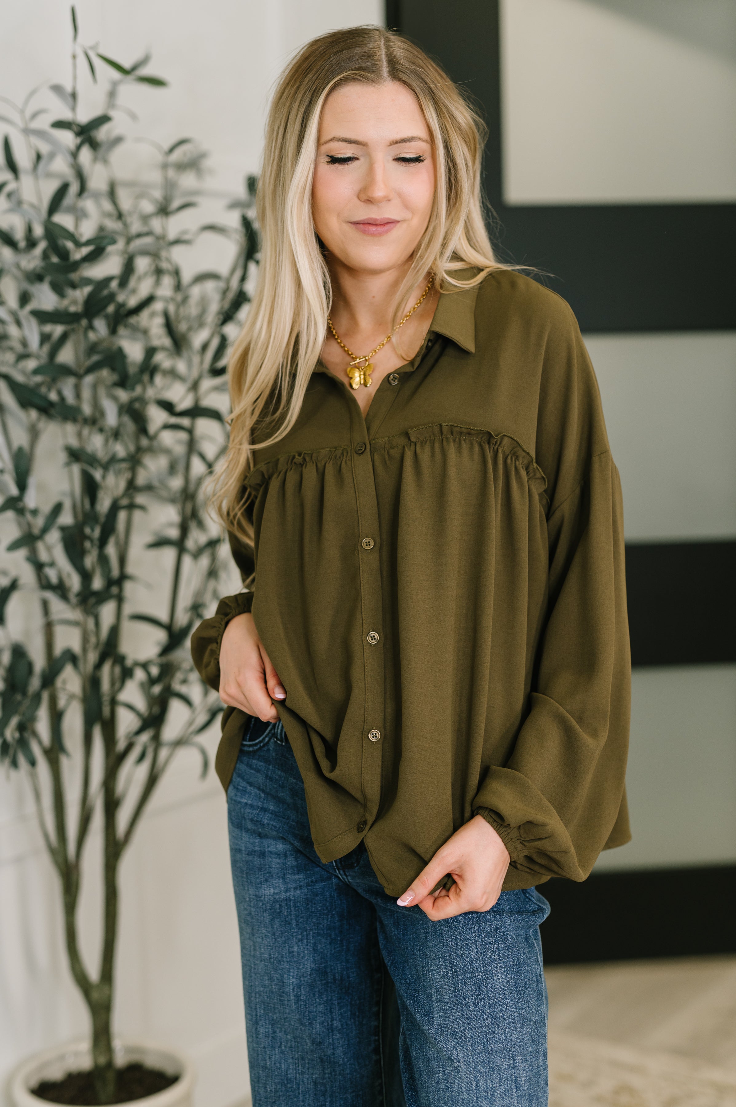 Woman wearing an olive green blouse and blue jeans indoors with a plant in the background