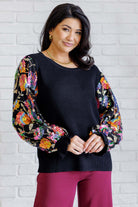 Woman wearing a black shirt top with colorful floral sleeves against a white brick wall.