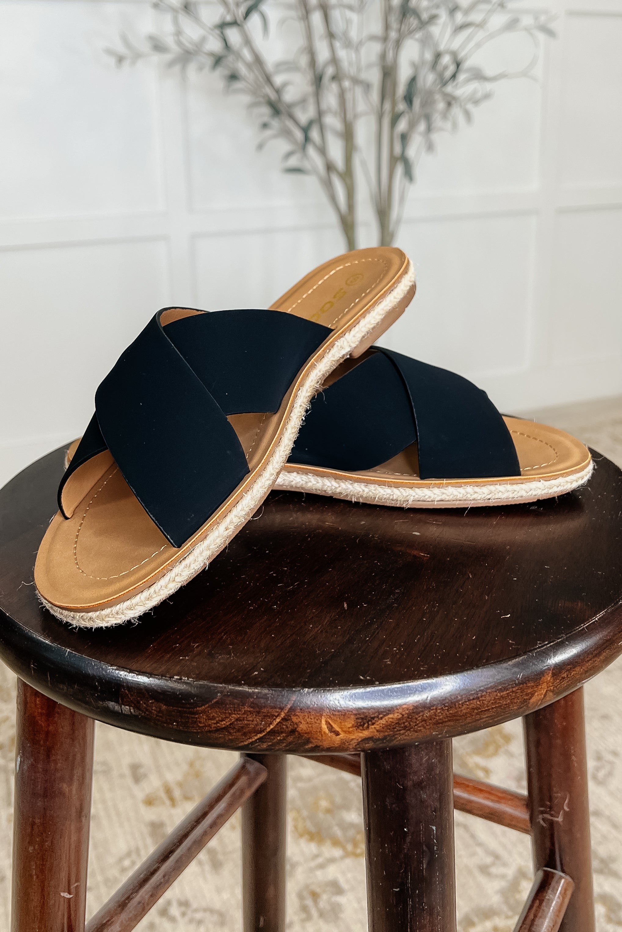 A pair of black slip-on sandals with crisscross straps and a braided twine detail on a wooden stool.