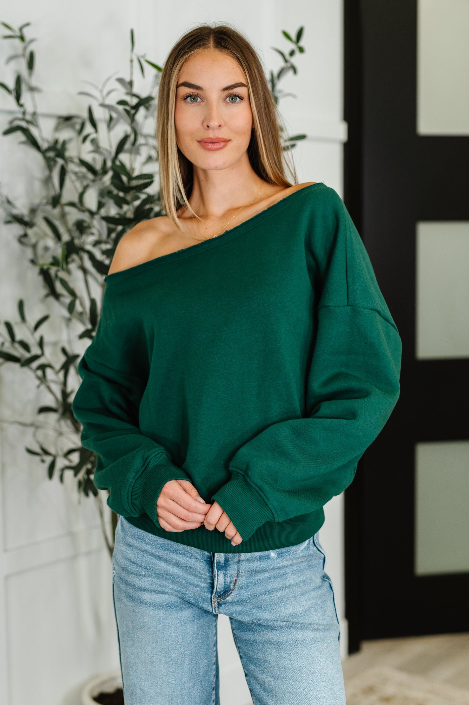 Woman wearing a green off-shoulder long sleeve shirt top and light blue jeans indoors.