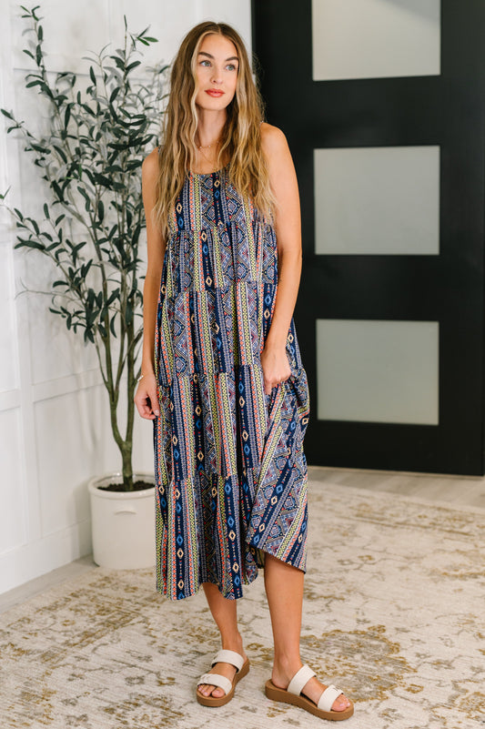 Woman wearing a patterned dress standing indoors with a plant and wall decor in the background.