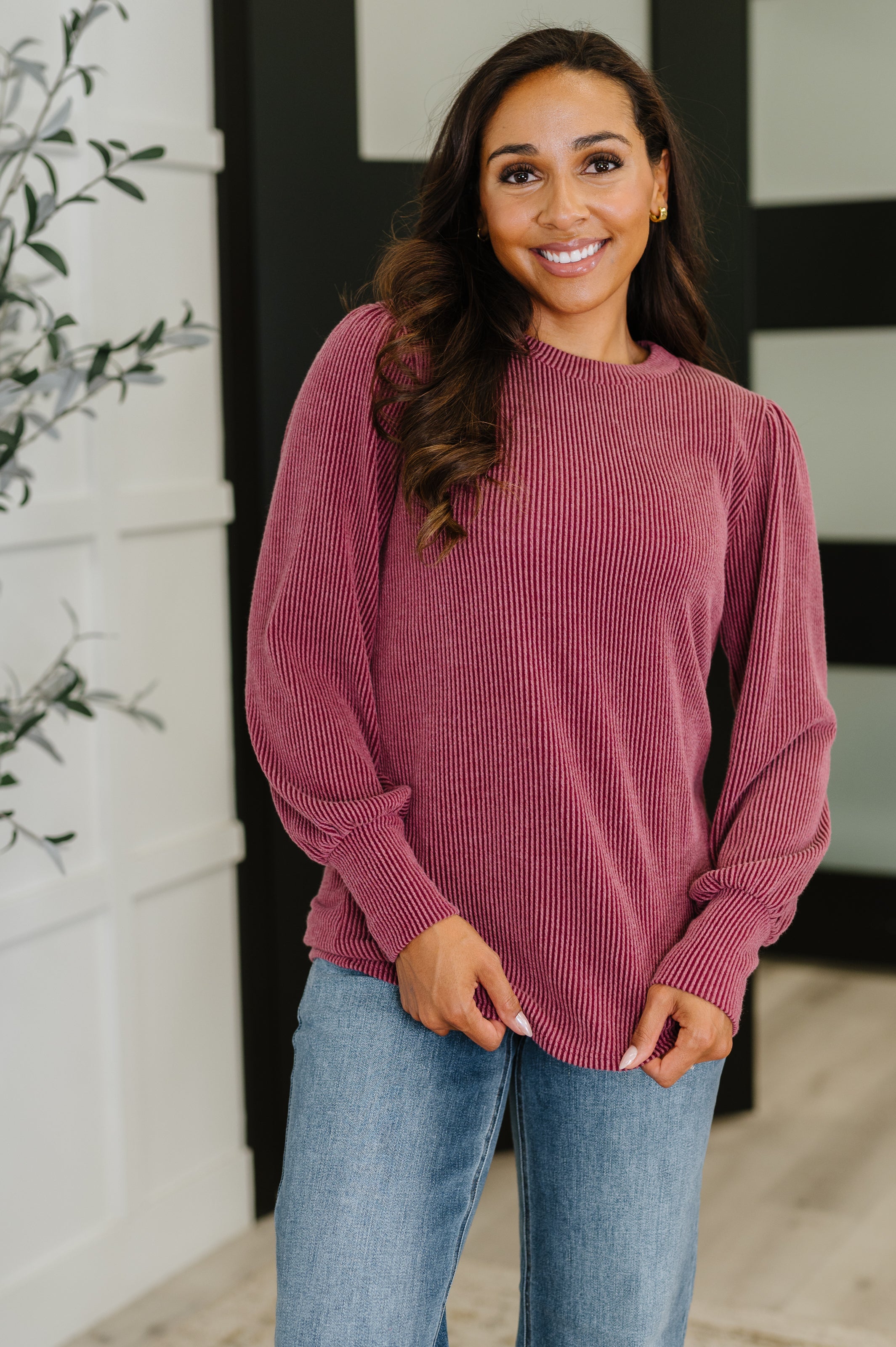 Woman wearing a maroon ribbed sweater and blue jeans standing indoors.