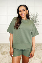 A woman wearing a light olive colored ribbed knit set with a banded round neckline top and matching shorts, featuring functional side pockets.