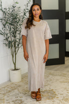 Woman wearing a beige dress standing indoors with a plant and decorative wall in the background