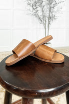 A pair of woman tan slip-on sandals displayed on a dark wooden stool, with a round toe and a single band design.