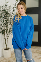 Woman wearing a blue sweatshirt and jeans standing indoors with a plant in the background