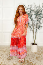 A woman wearing a red and orange floral maxi dress with a V-neckline and dolman sleeves, featuring a smocked elastic waistline, a sleeve cuff tie, and tassel detailing.