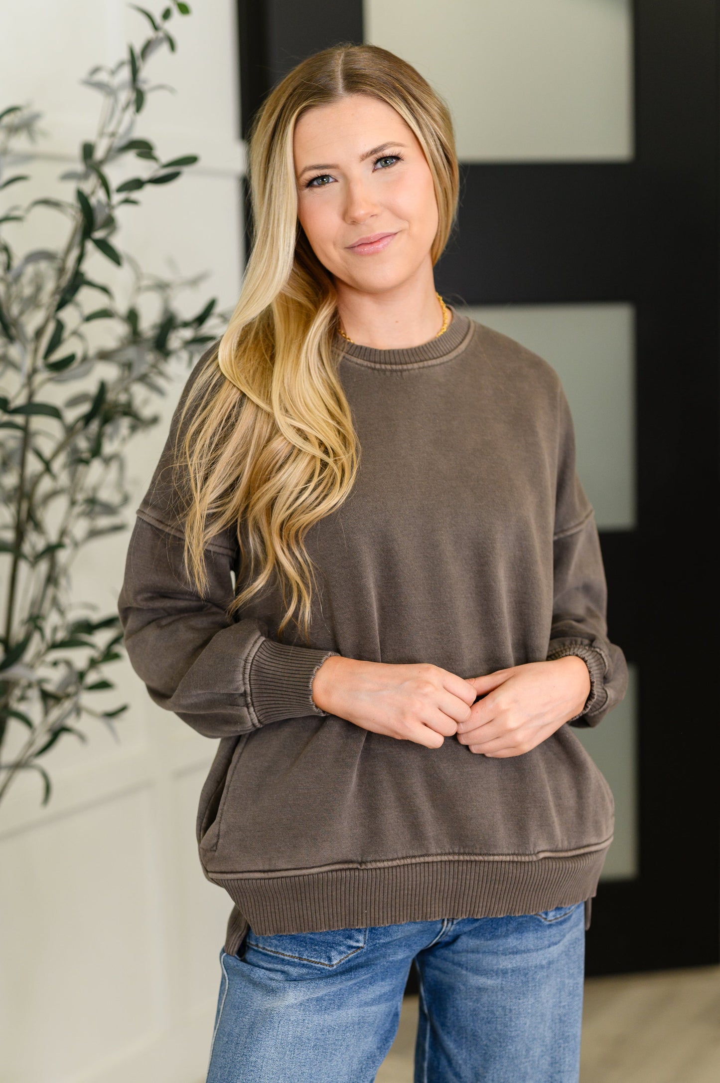Woman wearing a brown sweatshirt and blue jeans standing indoors with a blurred background