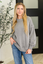Woman wearing a gray sweatshirt and blue jeans standing indoors with a plant in the background