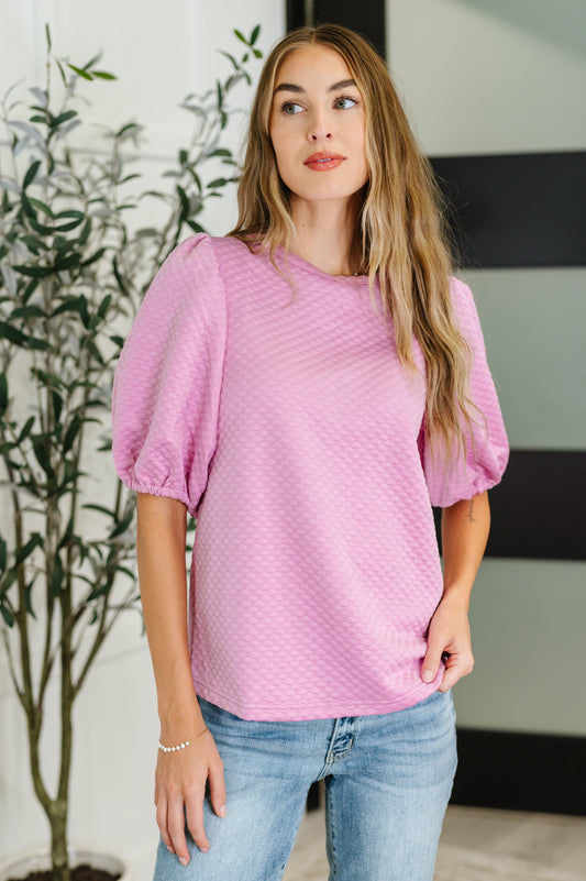 Woman wearing a pink textured top and blue jeans indoors with a plant in the background