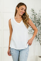 A woman wearing a sleeveless, v-neck, ribbed knit tank top in ivory color paired with blue ripped jeans.