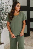 Woman wearing a green outfit standing indoors with a plant in the background