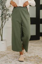 Woman wearing green pants and beige shoes in a room with a plant and decorative wall.