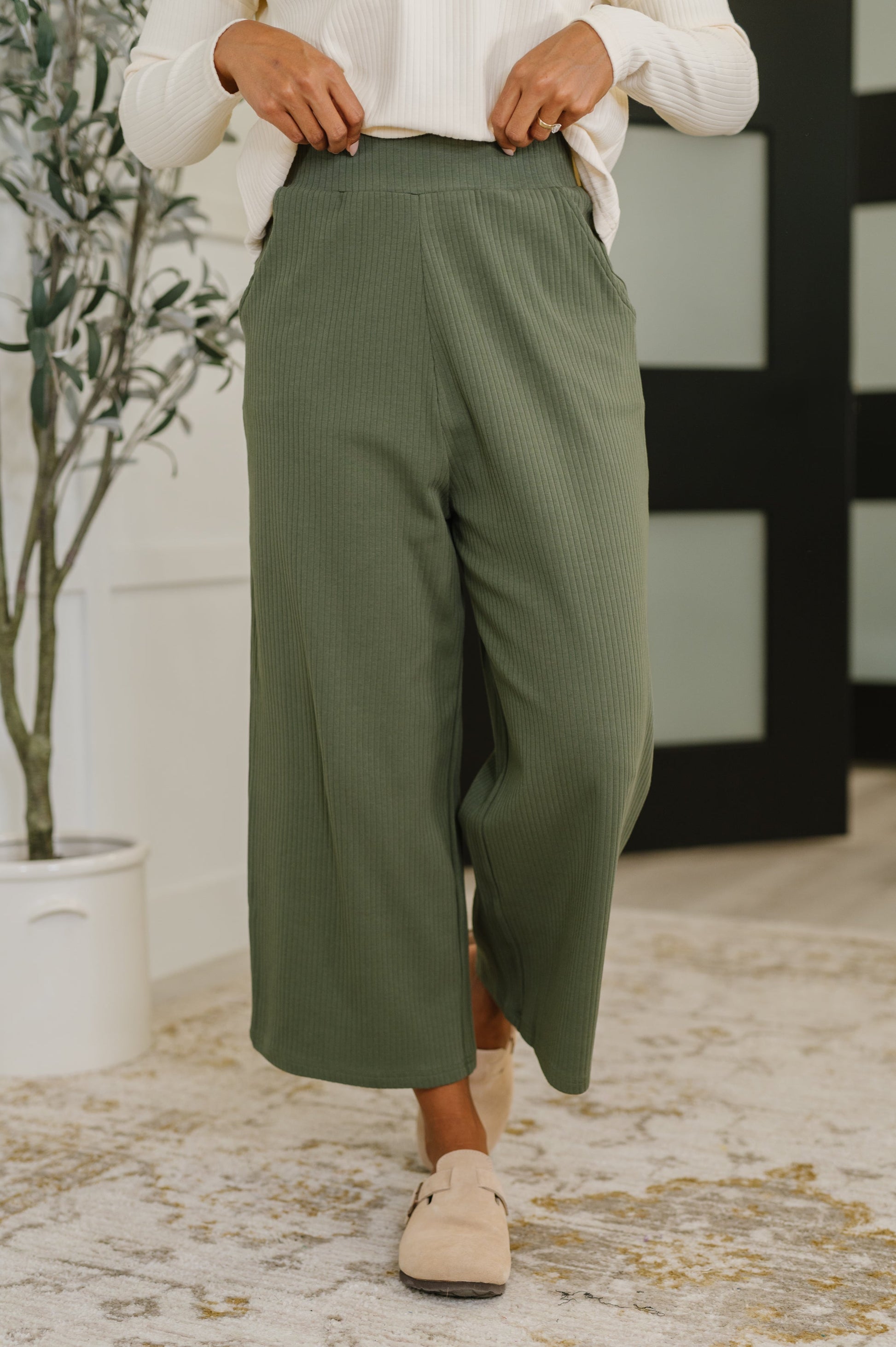 Woman wearing green pants and beige shoes in a room with a plant and decorative wall.