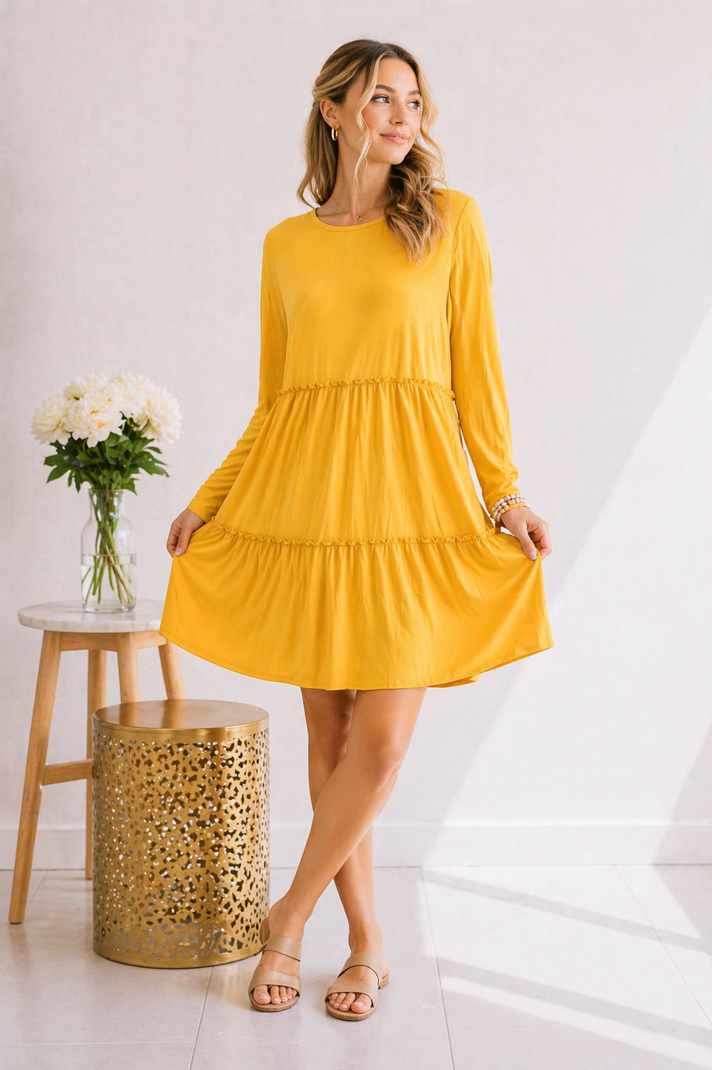 A woman wearing a long sleeve mustard dress with a tiered ruffle hem, standing next to a small table with flowers.