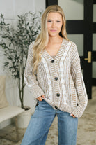 A beige cable knit cardigan with a deep V neckline and button-down closure, worn by a model with jeans.