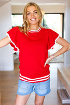 Woman wearing a red top with white trim and denim shorts indoors.