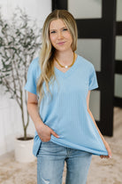 Woman wearing a light blue ribbed t-shirt and jeans with a blurred indoor background
