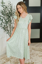 Woman wearing a light green floral dress indoors with a plant and wall decor in the background.