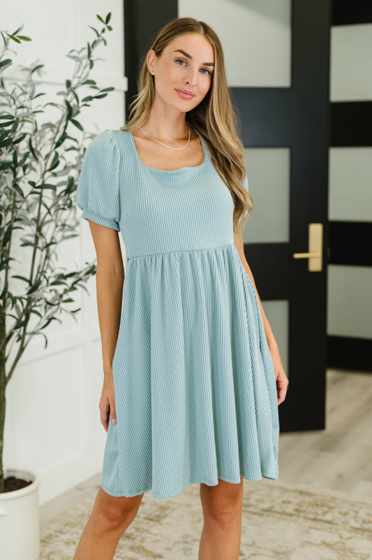 Woman wearing a light blue dress standing indoors with a plant and door in the background