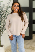 Woman wearing a beige shirt top and blue jeans standing indoors with a plant in the background