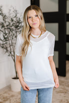 Woman wearing a sheer white top and jeans indoors with a plant in the background