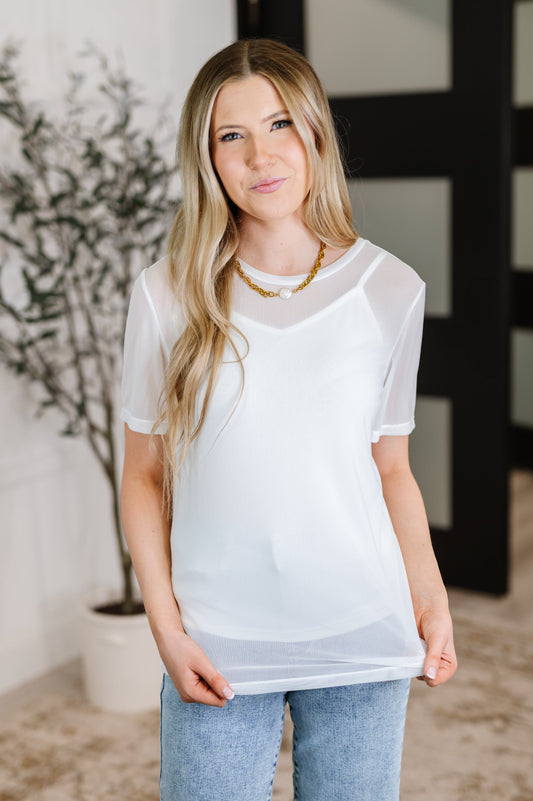 Woman wearing a sheer white top and jeans indoors with a plant in the background