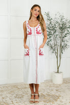 Woman wearing a white sleeveless scoop neck dress with red embroidery and functional patch pockets.