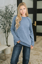 A woman wearing a dusty blue raglan sleeve sweater with ribbed cuffs and hem, featuring side slits.