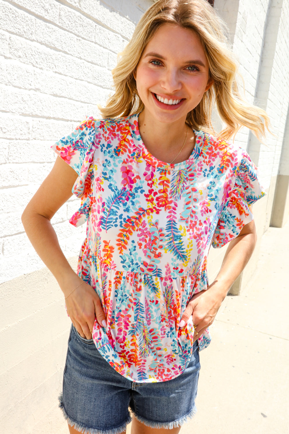 A woman wearing a multicolored floral leaf print short sleeve babydoll top with round neckline, paired with denim shorts.