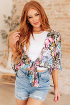 A woman wearing a multicolored floral front tie blouse with flutter sleeves and a scooped hem, paired with a white top and blue denim shorts.