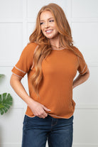 Woman wearing a rust-colored short-sleeve top and blue jeans against a white tiled wall.