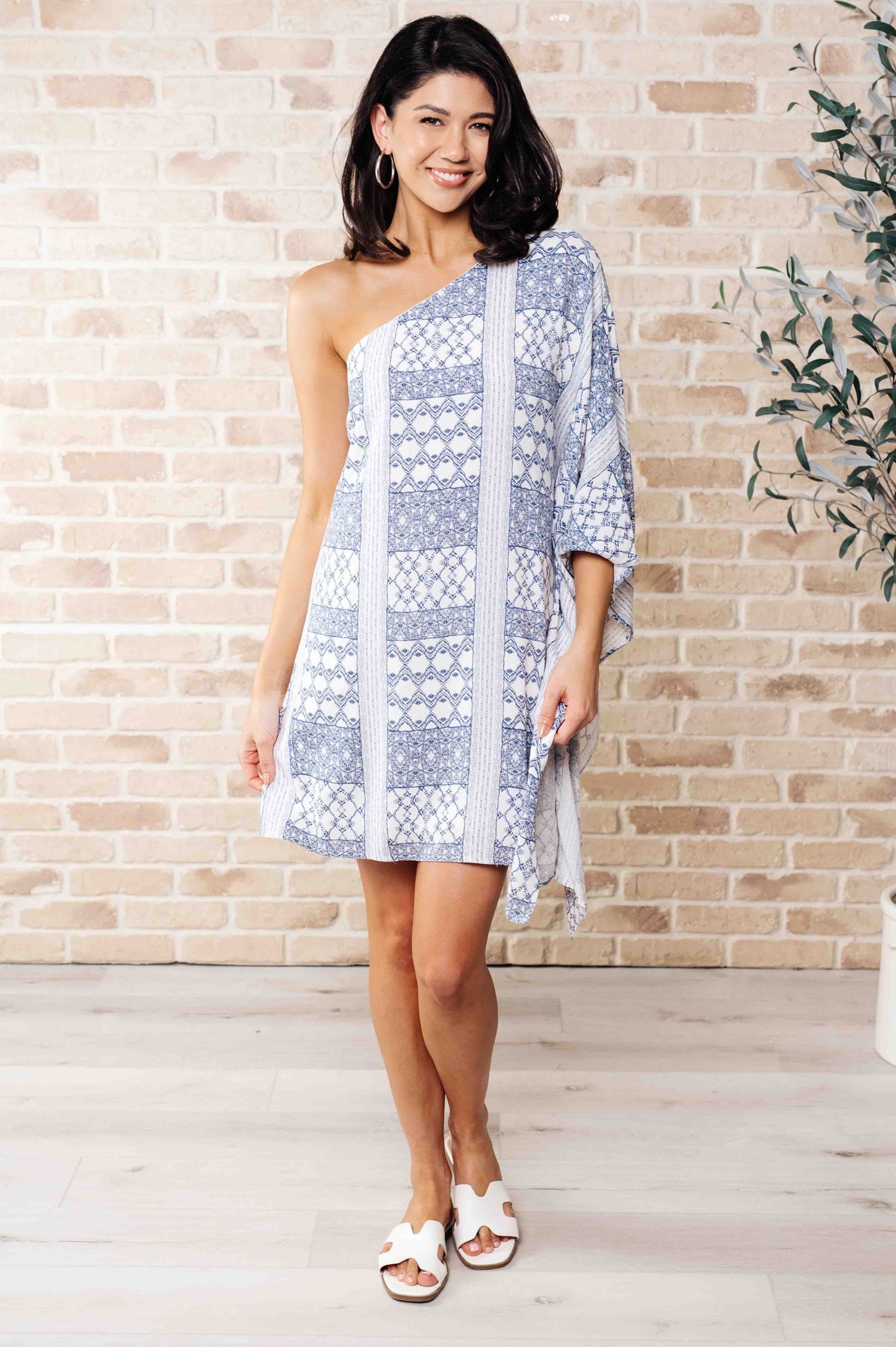 A woman wearing a blue and white one-shoulder mini dress with a geometric pattern, paired with white sandals.
