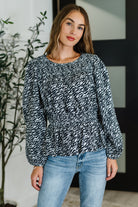 Woman wearing a black and white patterned blouse and blue jeans indoors.