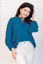 Woman wearing a blue blouse and white pants against a white brick wall.