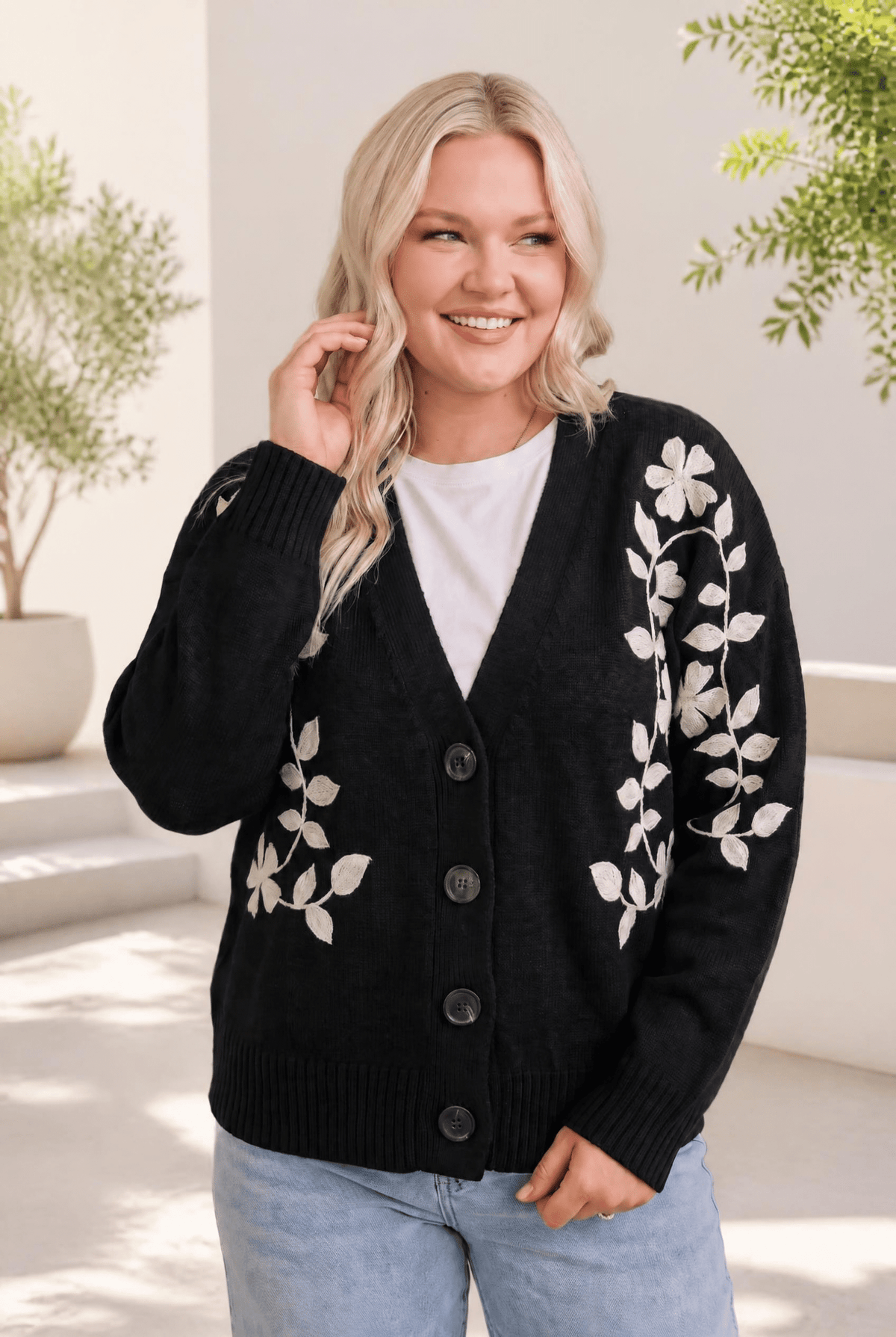 A woman wearing a black cardigan with cream floral embroidery, buttoned in front, with ribbed cuffs and a relaxed fit.