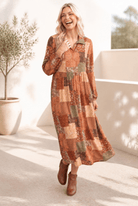 A woman wearing a patchwork midi dress with long sleeves, featuring a mix of browns, greens, and yellows, paired with brown ankle boots.