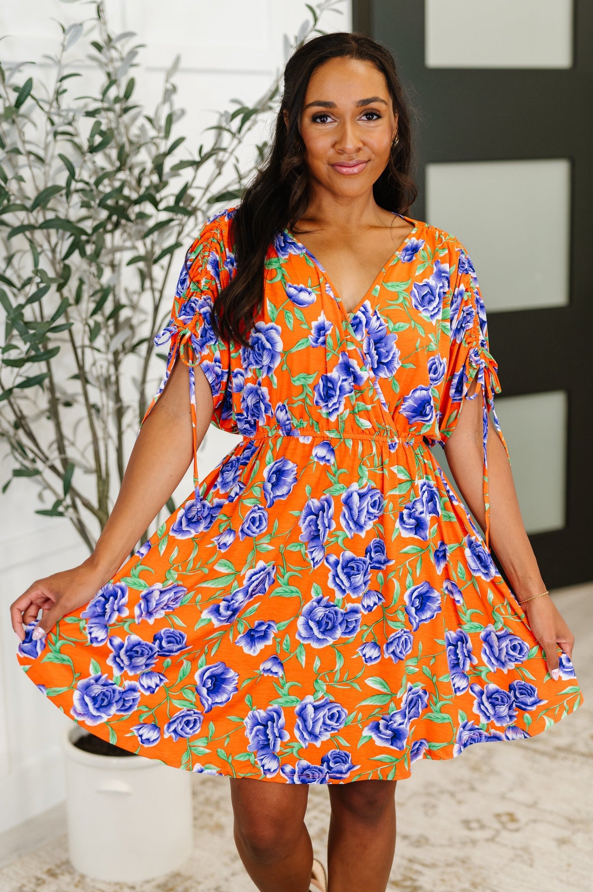 Woman wearing an orange floral dress standing indoors with a plant in the background