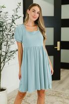 Woman wearing a light blue dress standing indoors with a plant and door in the background