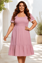 A plus-size midi dress in soft pink with a Swiss dot pattern, smocked bodice, and tiered hem.