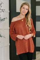 A woman wearing a rust-colored off-shoulder textured knit top with wide sleeves and ribbed cuffs.