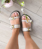 Women's White sandals with gold buckles worn on a light wooden floor with flowers.