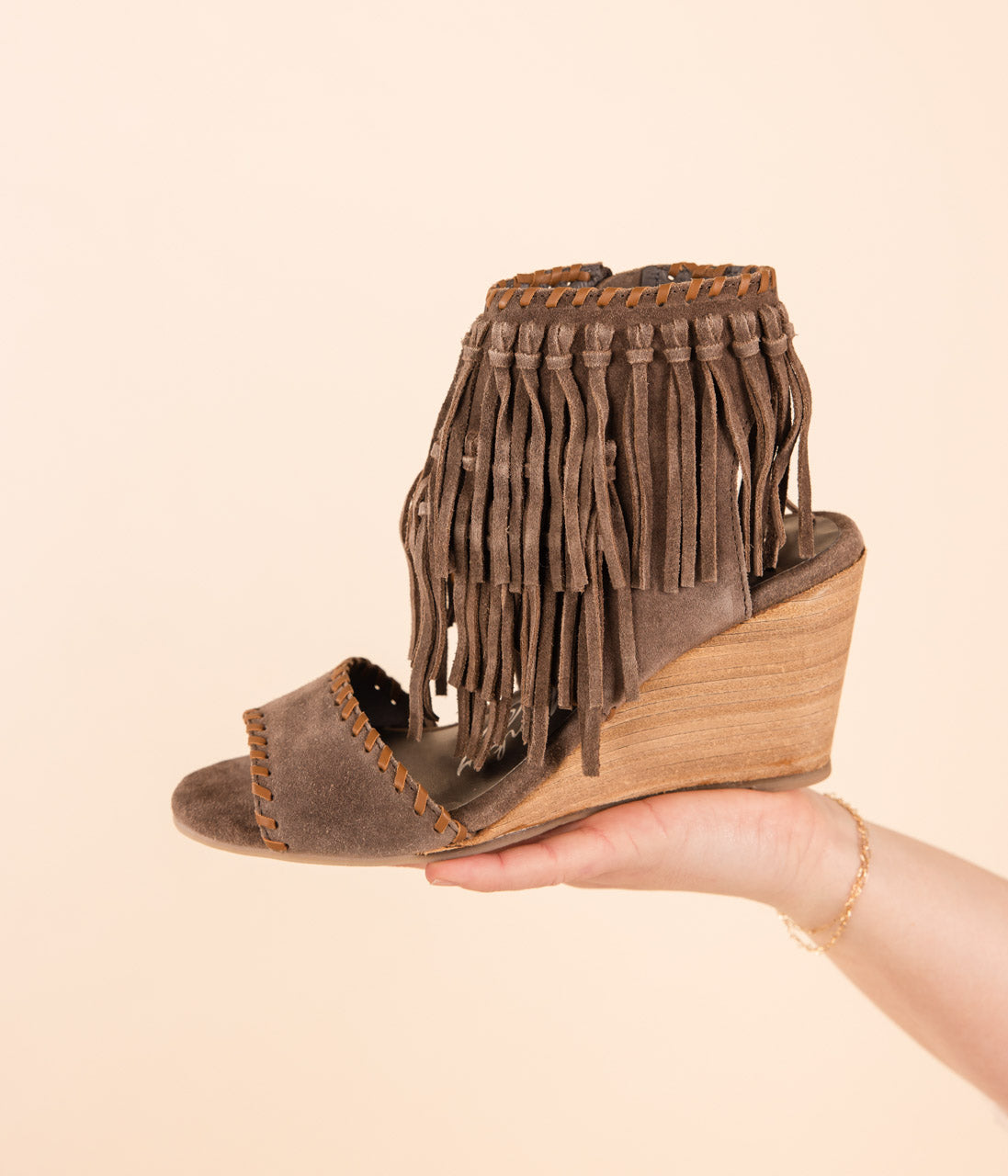 A taupe colored wedge shoe with fringe ankle detail, held in a person's hand against a light background.