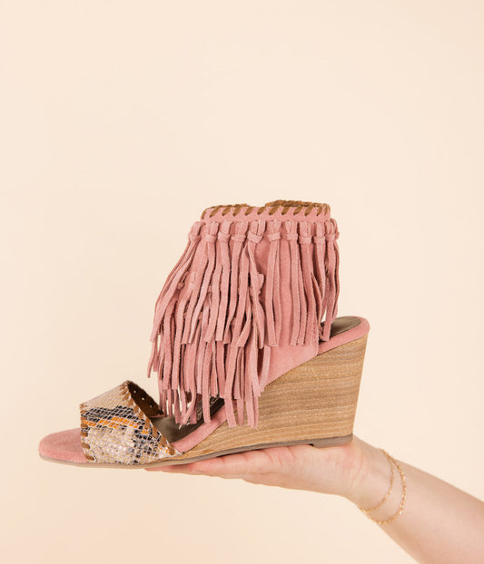 A blush pink fringe wedge sandal held in a hand against a plain background.